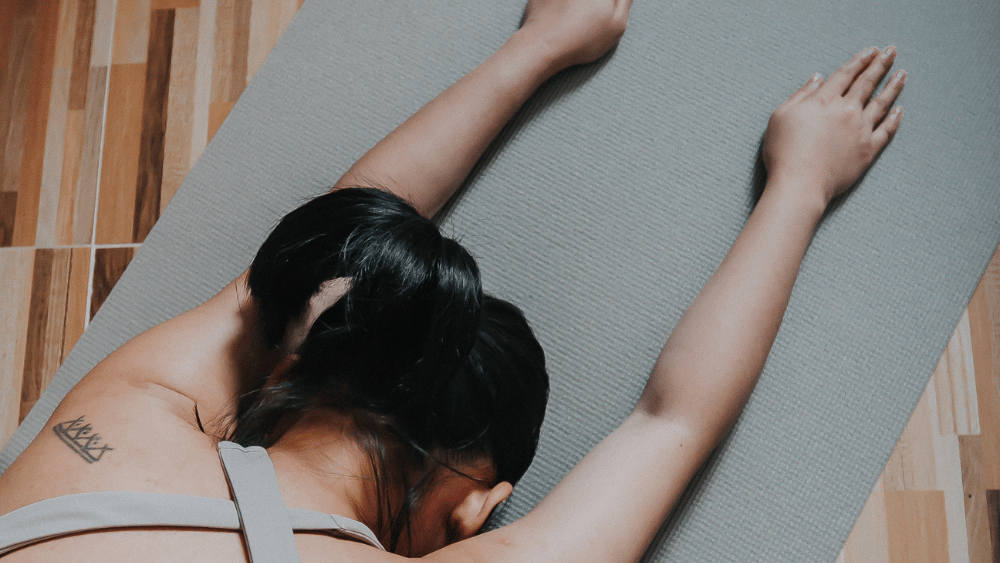 The Benefits of Yin Yoga for a Balanced Lifestyle 2 Person practicing Yin Yoga in Extended Child’s Pose on a gray yoga mat, with arms stretched forward and a tattoo visible on the shoulder blade.